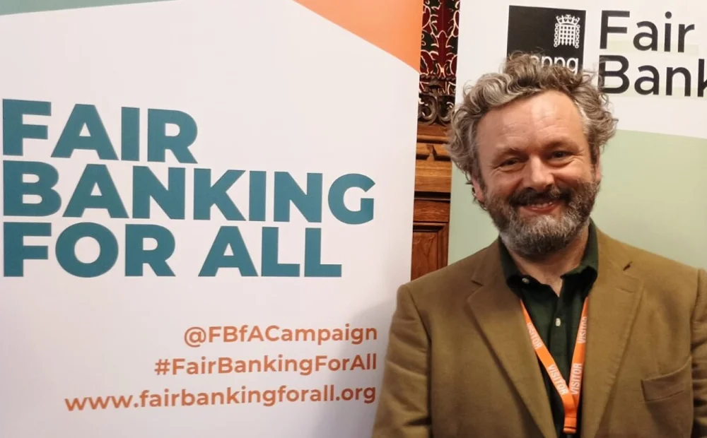 Michael Sheen standing in front of 'fair banking for all' sign Michael Sheen standing in front of 'fair banking for all' sign